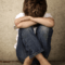 Young boy sitting near a dirty wall with his feet pulled up, arms crossed over them, and head bent down