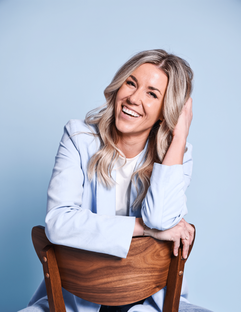 Smiling woman in a light blue blazer sitting on a wooden chair, set against a pale blue background.